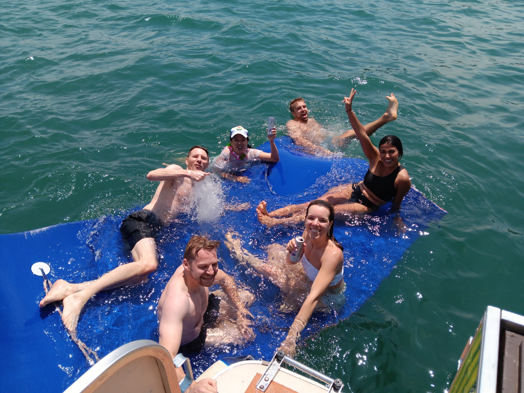 Friends floating and waving on a big blue water mat beside the boat
